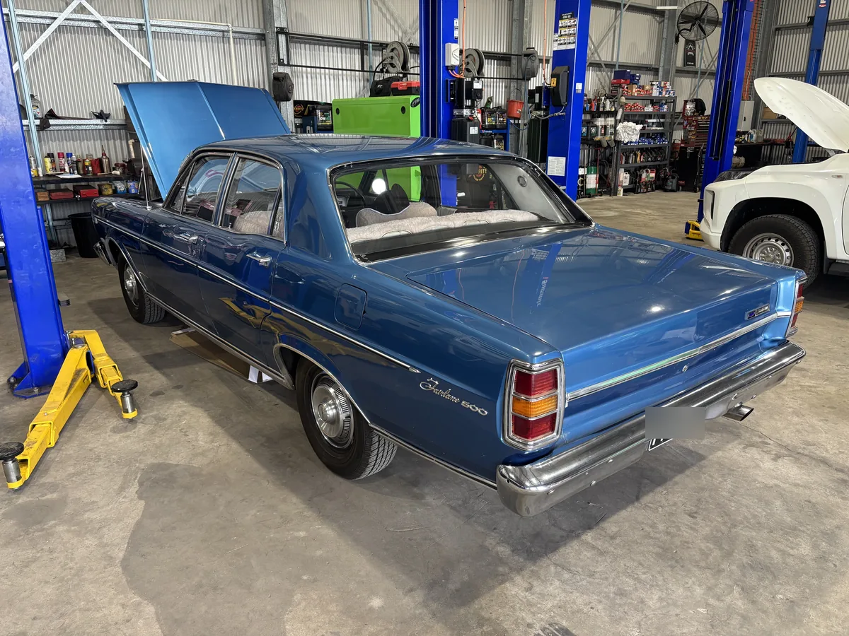A blue Ford Fairlane 500 parked in the workshop between two blue hoists with its bonnet open.