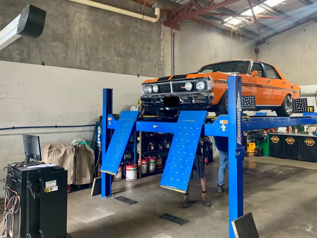 An orange classic car raised on a blue hoist in the workshop.