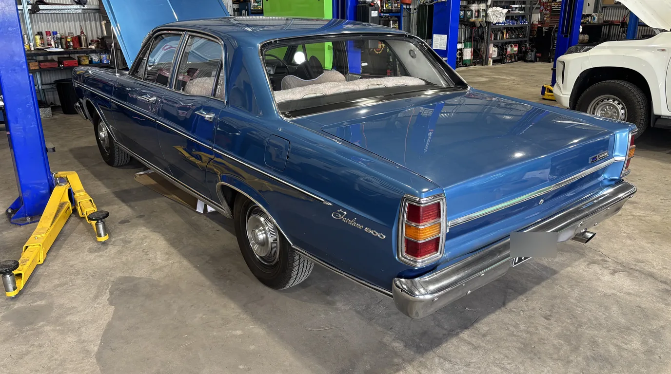 A blue Ford Fairlane 500 parked in the workshop between two blue hoists with its bonnet open.