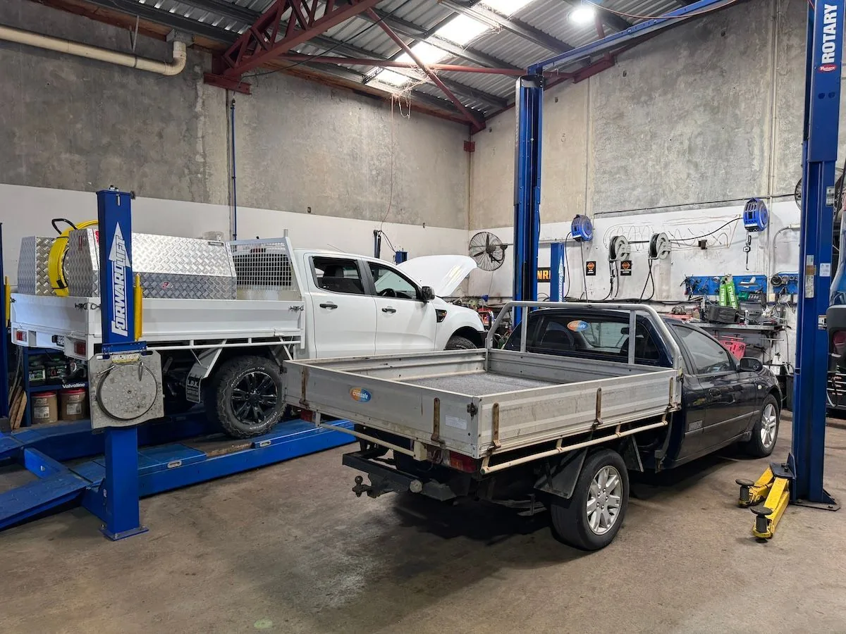 Two utes raised on blue hoists in the workshop, one with its bonnet open.