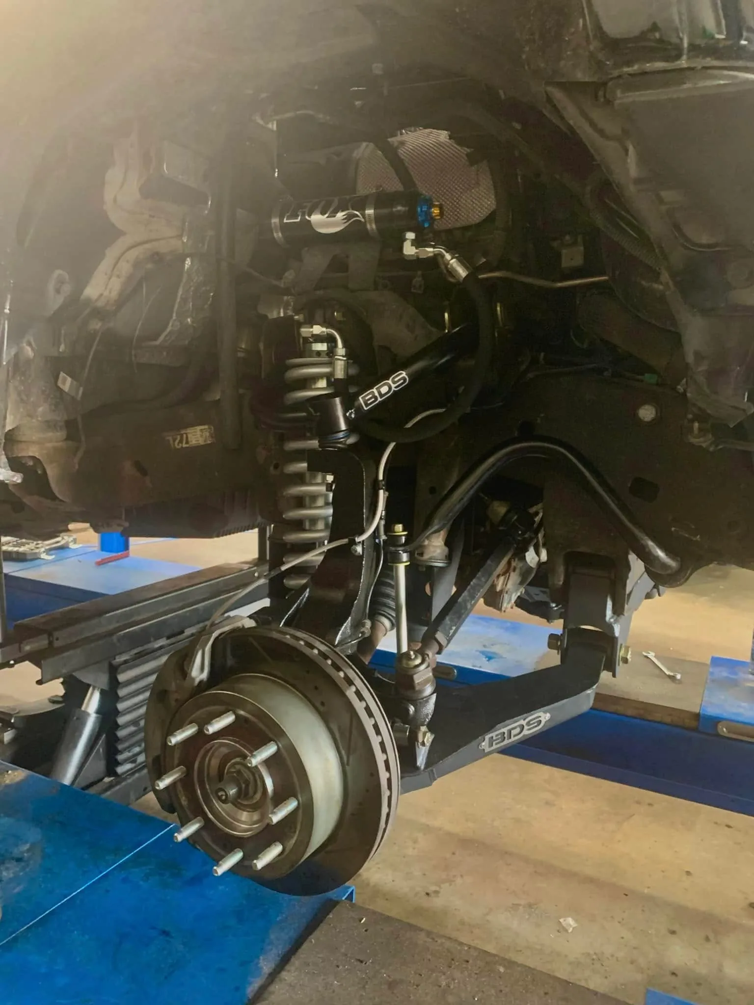 A vehicle raised on a hoist with a coil-over shock absorber and brake disc exposed at the front wheel.